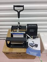 Front view of the Bettersun heat press machine set on top of its original cardboard packaging. The unit is black, with digital temperature controls visible on the side. User manual placed next to the heat press.