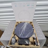 Robot vacuum device in original packaging showing main unit still wrapped in plastic along with other accessories.