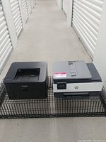 Photo showing a Canon black laser printer and an HP gray/black laser printer side by side on a wire rack in a hallway with storage unit doors.