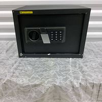 Front view of small black electronic safe featuring digital keypad lock and manual turn handle on a lace fabric covered surface.
