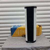 Front view of the ceramic heater standing on a table with its original box behind it, showing the white and black design and vents.
