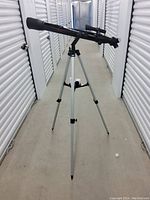 Side view of the black refractor telescope mounted on an adjustable silver tripod in a storage hallway.