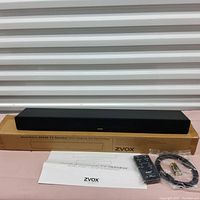 Zvox AccuVoice AV355 center speaker placed on its original cardboard box with remote control sealed in plastic, two AAA batteries, coiled audio cable, and folded instruction manual all arranged neatly for display.
