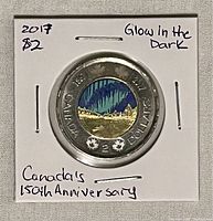 2017 Canadian $2 coin in white cardboard holder with notes "2017 $2", "Glow In The Dark", "Canada's 150th Anniversary." Coin shows full bimetallic design with outer silver ring and colorful glowing center.