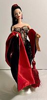 Front view of Barbie doll wearing beaded dress and red/black shawl, holding a tag reading 'Dans Work Collection'. Doll on clear display stand.