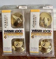 Front view of two sealed Weiser lock packages showing brass-colored deadbolt locks and thumb turns visible through clear plastic packaging.