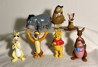 Photo showing the collection of 8 Winnie the Pooh character figures arranged standing, visible characters include Pooh, Tigger, Piglet in native attire, Eeyore, Owl, Rabbit, Kanga with Roo.