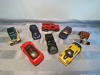 Eight Hot Wheels model cars arranged on fabric surface showing top and side views. Various colors and styles visible, including sporty and classic models.