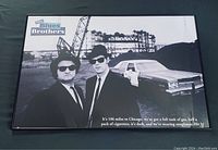 Full view of the framed black and white Blues Brothers poster showing John Belushi and Dan Aykroyd in suits, sunglasses, and hats, with car and industrial background.
