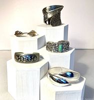 Lot of five cuff bracelets in various metal tones displayed on white block stands showing different angles and designs.