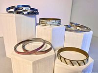 View of 8 shell inlaid bangle bracelets displayed on white hexagonal stands, showing color variety and design differences.
