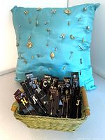 Teal satin pillow with assorted metallic letter and leaf stick pins attached, additional pins are placed upright on black card mounts inside a woven basket in front of the pillow.