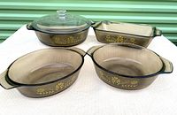 Five-piece vintage Glasbake bakeware set includes three oval/round casseroles, one rectangular loaf pan, and one large clear lid. All smoky glass with yellow primrose floral design.