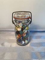Photo of vintage Wheaton Glass Co. glass jar filled with assorted vintage marbles showing clear glass, hinged lid with metal clasp, and rubber gasket