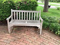 Full view of wooden outdoor bench on brick patio, showing entire bench with slatted back and seat, armrests, and weathered condition.