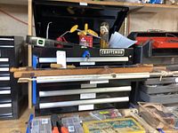 Front view of black Craftsman tool chest with five drawers, lift-off top raised, and key inserted in lock. Chest placed on a basement shelf with various tools and items around, including wooden objects resting on top.
