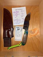 Knife and leather sheath laid on wooden shelf with seller note and MaxSold tag
