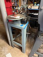 Full side view of Delta scroll saw mounted on grey metal stand with power motor and electrical cords visible. Floor and basement setting visible in background.