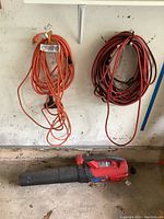 Two heavy-duty extension cords, one orange and one dark red, hanging on a wall with Toro PowerJet 1700 electric leaf blower in front on floor.