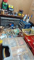 View of assorted hardware items stored in plastic organizer cases, bins, and jars against pegboard wall.