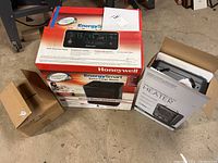 Photo of three boxed items: Honeywell EnergySmart infrared heater box, electric utility heater box, and ceiling exhaust fan box on basement floor.
