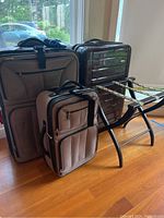 Photo showing three pieces of luggage and a folding luggage rack in a room with wooden floor and windows.