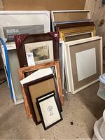 Photo showing a collection of various sized empty picture frames stacked upright including large white IKEA RIBBA frame, brown wooden frames, gold frame, and smaller black frames.