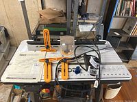 Full view of Ryobi router table with orange adjustable fence, power cord visible, router under table, manual on table surface.