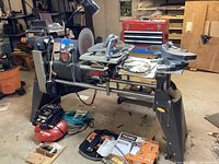 ShopSmith woodworking system mounted on black metal motor stand with 6 inch belt sander, 12 inch disc sander, circular saw, and accessories. Various tools and manuals placed nearby.