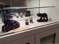 Wide shot of all four stone figurines and eggs inside a glass cabinet: blue tiger on wooden base, dark elephant on wooden base, two stone eggs each on their own wooden stands.