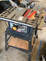 Front side view of Ryobi 10 inch table saw on metal stand with wooden shelf, showing saw surface, push stick, and stand structure.
