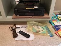 TEAC CD changer unit on shelf with remote control, manual, and power cord visible in front. Additional plastic bag with accessories also visible.