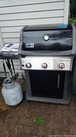 Front view of Weber Spirit grill with control knobs, side shelf, attached propane tank and utensils hanging