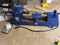 Full side view of the blue Record Drillmaster Power wood turning lathe showing motor, spindle chuck, tool rest, and tailstock on concrete floor.