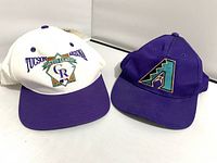 Two baseball caps side by side, purple AZ Diamondbacks cap and off-white Cactus League cap with purple brim and logos
