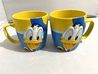 Front-facing view of two ceramic Donald Duck mugs side by side, showing the full face of the character and the bright yellow handles and rims.