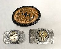 Front view of three belt buckles: an oval tooled leather buckle with leaf design, a silver-tone buckle with an oval stone, and a silver-tone buckle with a gold tone rooster.