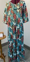Full view of a vintage muumuu dress with a turquoise blue base and large tropical floral prints, laid on a dress form without display stands.