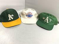 Three vintage baseball caps displayed side by side on a white surface: two Oakland A's caps in green and yellow and one white Chevrolet promotional cap.