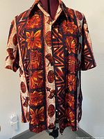 Front view of vintage Hawaiian shirt showing bold floral and tribal print pattern in orange, red, brown and cream colors, medium size.