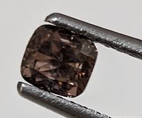 Close-up image of one cushion cut brown diamond held by tweezers, showing facets and color tone.