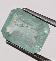 Close-up image of green emerald gemstone held by tweezers showing emerald cut and internal inclusions.