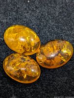 Three oval polished amber gemstones showing natural internal inclusions and rich golden to honey yellow tones placed on a black textured background.