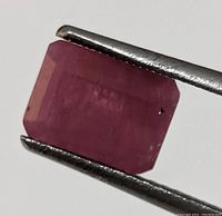 Close-up of one rectangular step cut genuine ruby gemstone held in metal tweezers showing surface and clarity details on white background.