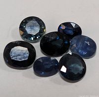 Six faceted oval shaped sapphires in various shades of blue and dark blue scattered on white background.