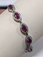 Close-up of the bracelet showing oval red rubies surrounded by clear cubic zirconia stones in sterling silver settings, displayed on a cream jewelry holder.