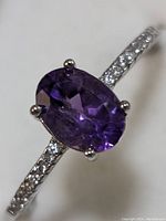 Close-up of sterling silver ring with oval faceted amethyst, prong set, showing small clear stones on band.