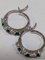 Close-up view of one sterling silver hoop earring showing green and clear prong-set stones.