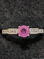 Close-up of the ring showing the round faceted pink ruby center stone with prong setting and small clear stones on the band against black background.