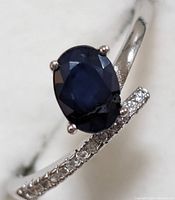 Close-up image of the oval dark blue faceted sapphire set on sterling silver ring band with small clear accent stones on one side.